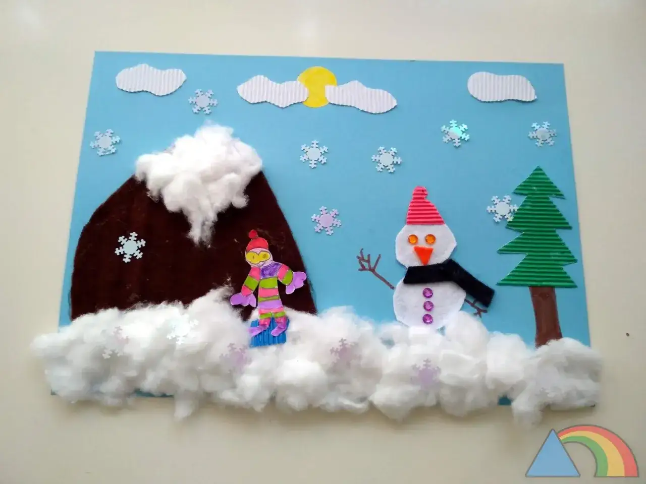 Manualidades invierno: un mu&ntilde;eco de nieve saluda cerca de un &aacute;rbol y un snowboarder desciende una monta&ntilde;a nevada bajo un sol amarillo.
