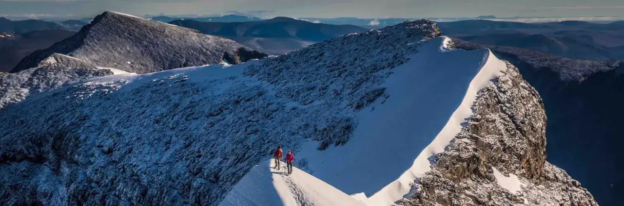 Två vandrare på en snötäckt bergskam i svenska fjällen. Majestätiska berg i bakgrunden.