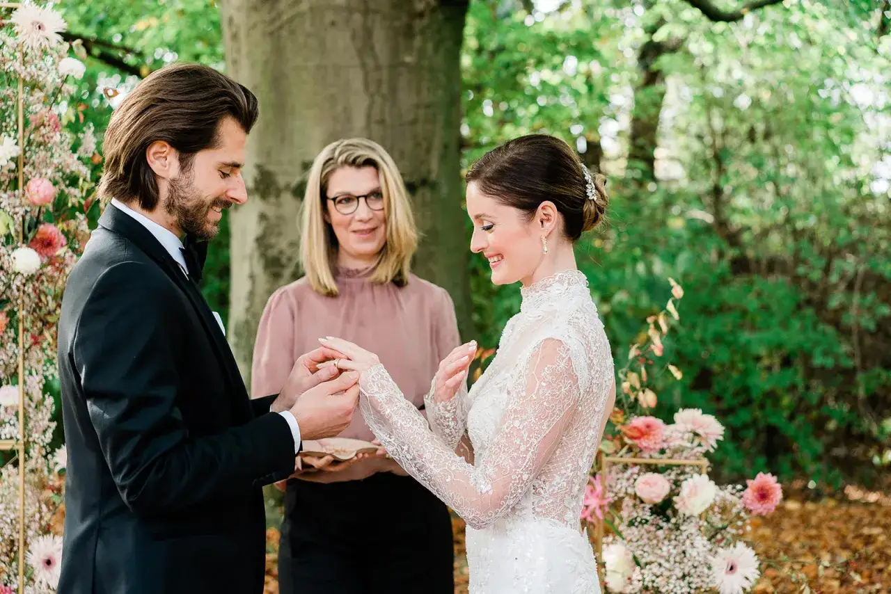 Ein Mann im Anzug steckt seiner Braut, die ein Spitzenkleid trägt, den Ring an. Eine Frau im rosa Oberteil beobachtet die emotionale Rede beim Standesamt.