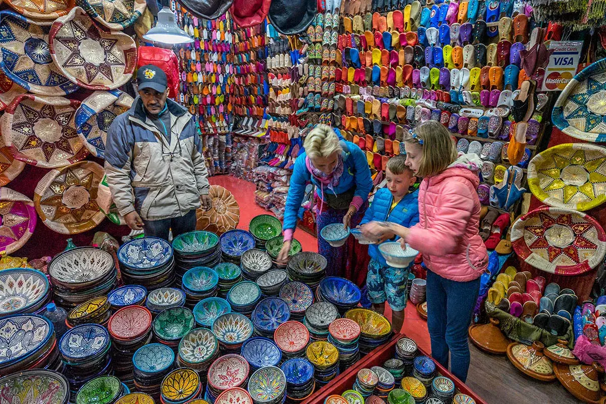 Kolorowy bazar podczas maroko wycieczka objazdowa. Sprzedawca prezentuje ręcznie malowane misy, a turyści oglądają kolorowe kapcie.