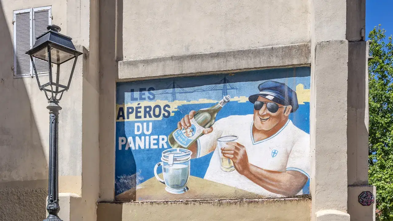 Ein Mann mit Sonnenbrille und Kappe schenkt in der **Marseille Altstadt** Aperitif aus. Das Wandgemälde