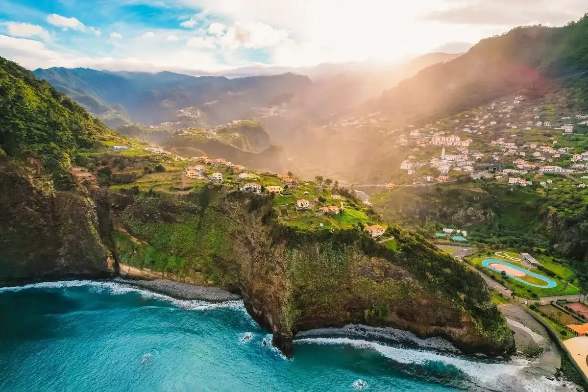Vue a&eacute;rienne de falaises verdoyantes plongeant dans l'oc&eacute;an bleu, avec un village pittoresque accroch&eacute; &agrave; flanc de colline. Un d&eacute;cor idyllique pour savoir quand partir &agrave; Mad&egrave;re.