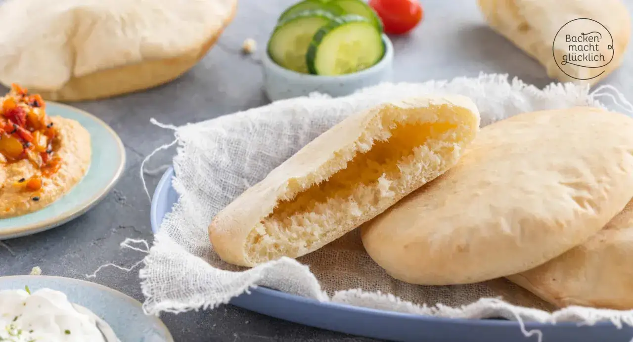 Frisches, aufgegangenes pita brot liegt auf einem blauen Teller, bereit zum F&uuml;llen. Daneben Hummus und Gurkenscheiben.