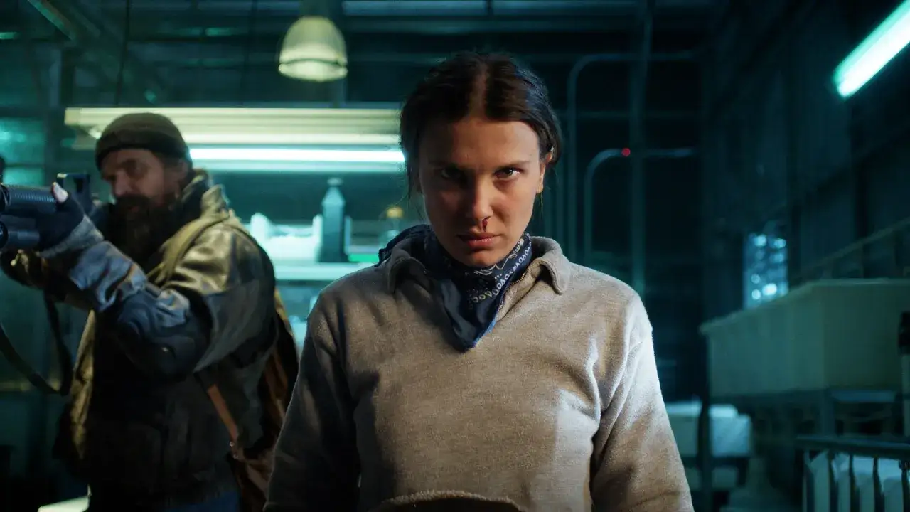 Nastka z Stranger Things, z bandaną na szyi i lekko zakrwawionym nosem, patrzy groźnie. W tle mężczyzna z bronią.