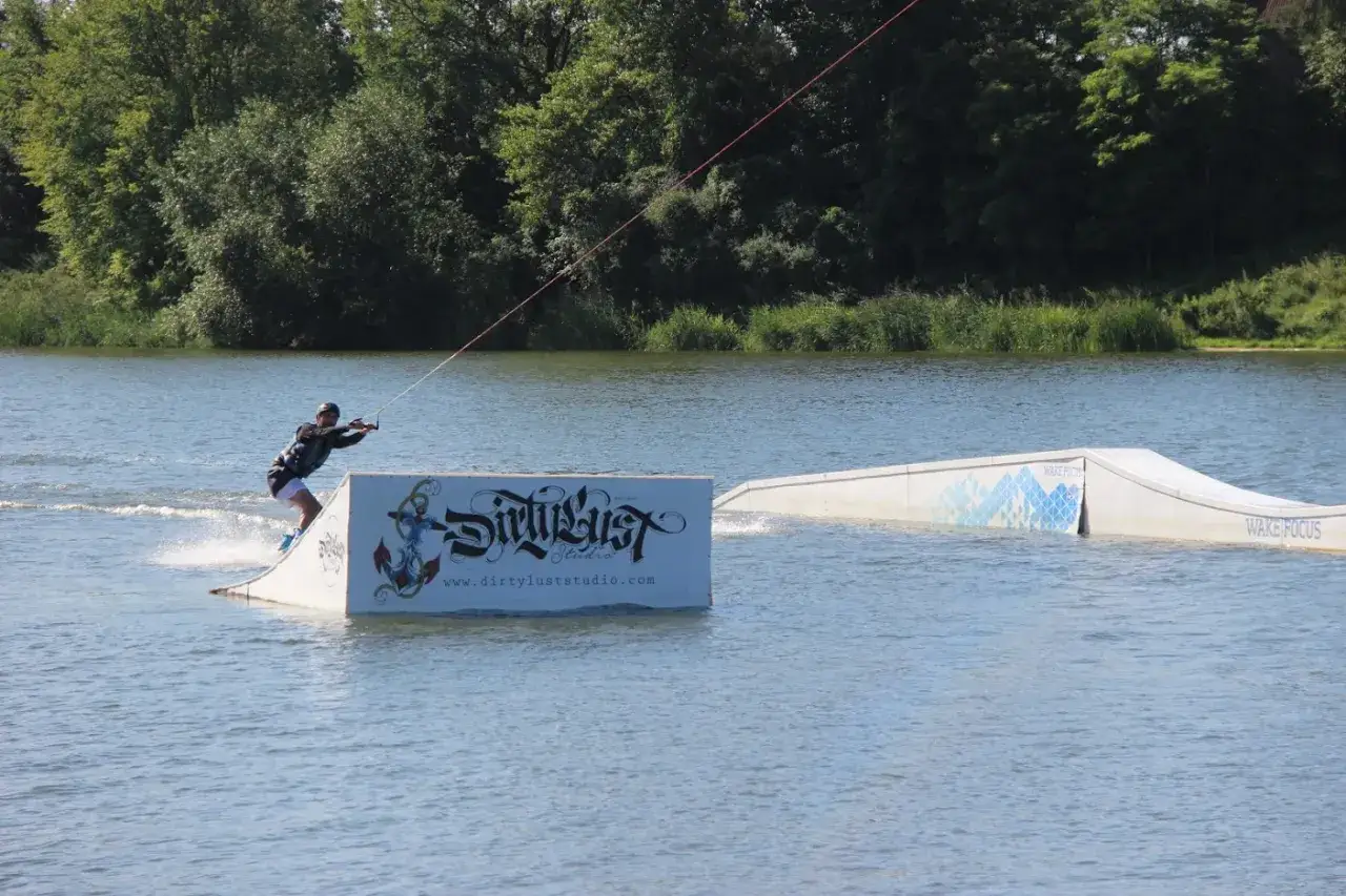 Osoba na desce, ciągnięta przez linę, wykonuje trik na wodzie. To właśnie jest wakeboarding, co to za sport?