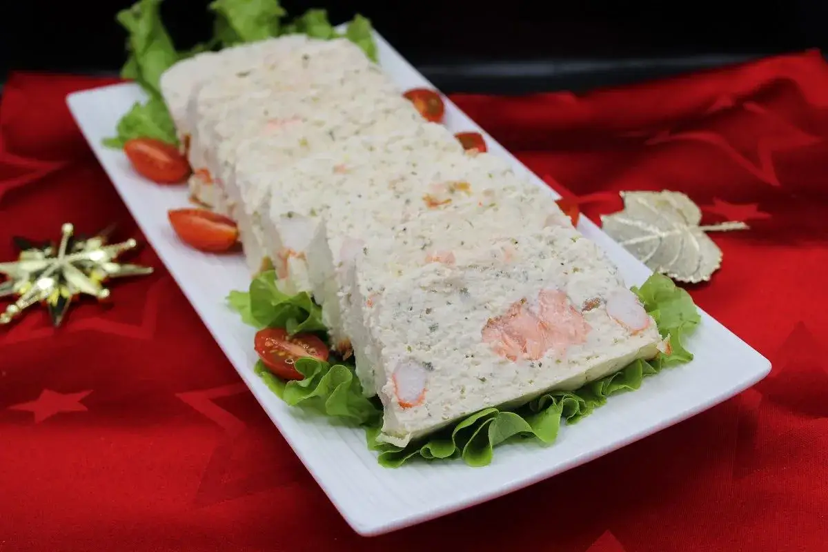 Tranche de terrine de poisson festive, garnie de crevettes et saumon, sur un lit de laitue et tomates cerises.