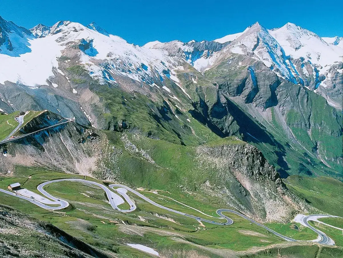 Serpentinenstraße schlängelt sich durch grüne Hänge zu schneebedeckten Gipfeln. Ein perfekter Roadtrip Alpen.