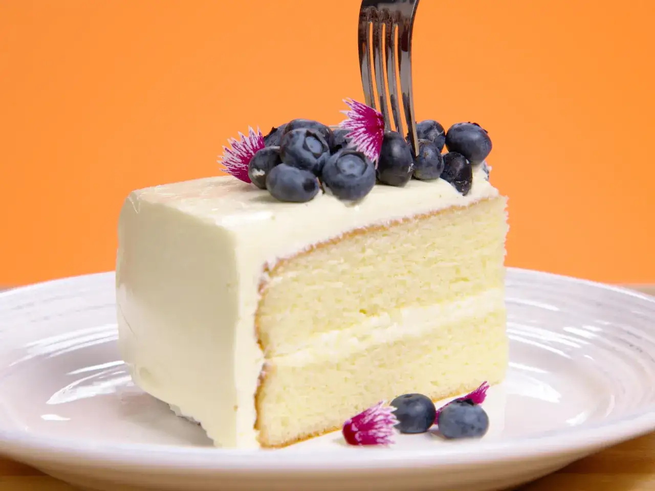 Tranche de g&acirc;teau moelleux, napp&eacute;e d'une onctueuse cr&egrave;me au beurre, d&eacute;cor&eacute;e de myrtilles et de fleurs. Une fourchette s'appr&ecirc;te &agrave; d&eacute;guster ce d&eacute;lice.