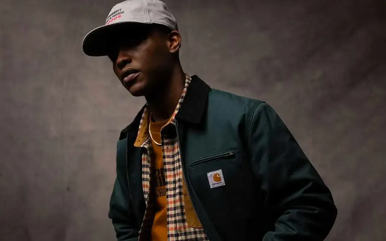 Un homme porte une veste verte foncée, une chemise à carreaux et un t-shirt jaune, incarnant le style workwear homme.