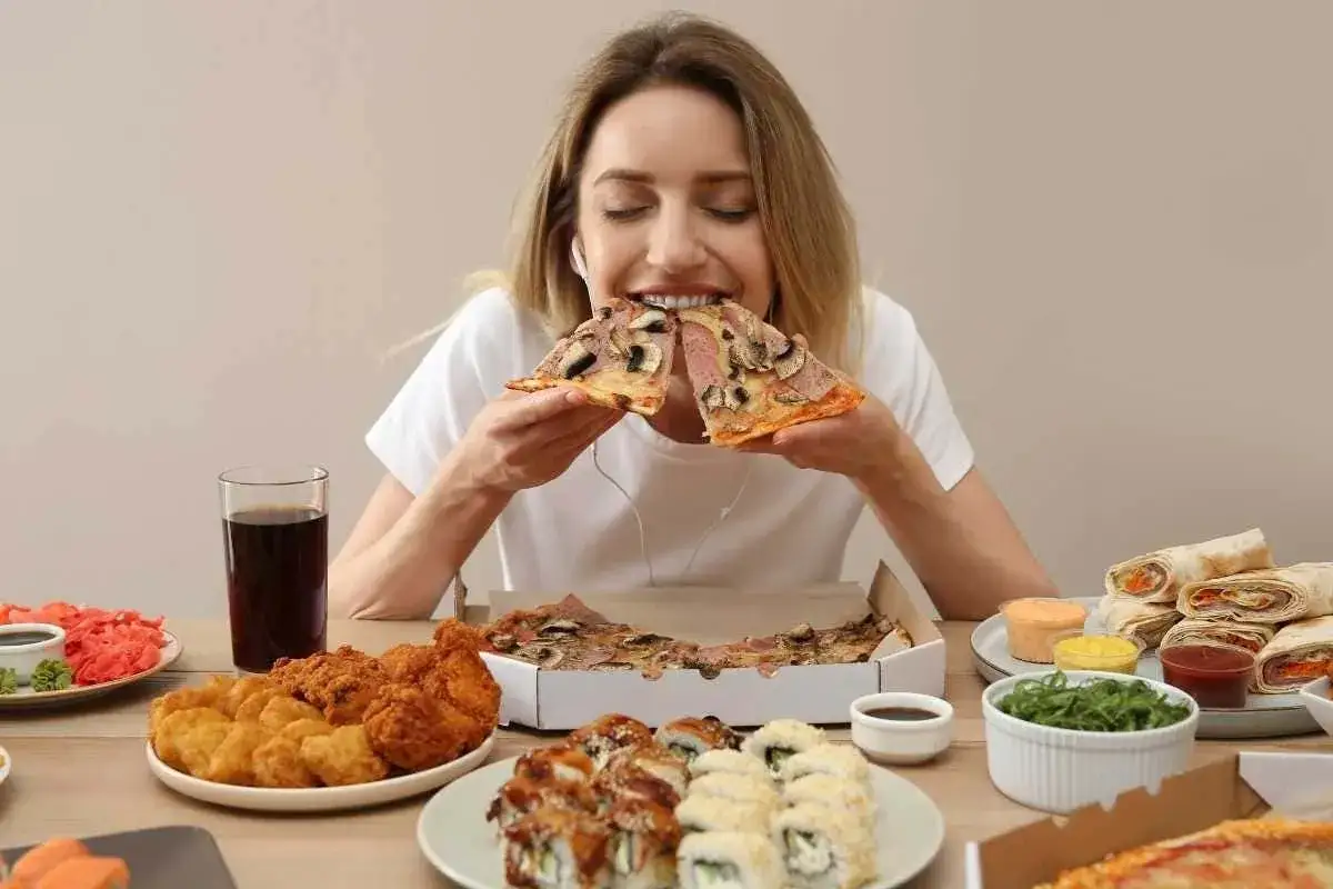 Donna mangia pizza, circondata da cibo spazzatura. Forse cerca rimedi per il mal di stomaco dopo questa abbuffata.