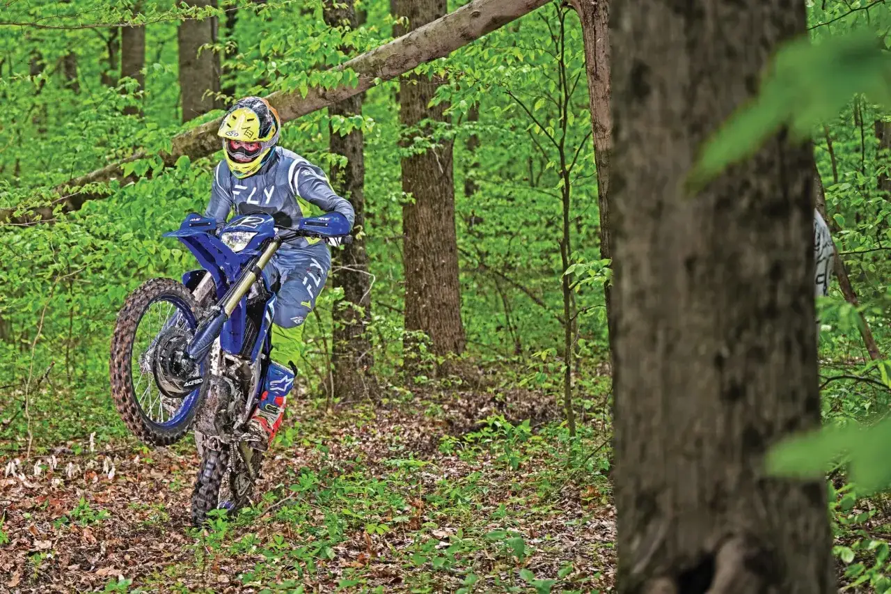 Piloto en una Yamaha WR450F azul levanta la rueda delantera en un sendero boscoso.