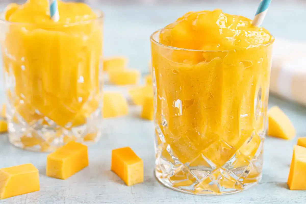 Erfrischendes Slush Eis selber machen: Gelbe Fruchtstücke und zwei Gläser mit leuchtend gelbem Slush, garniert mit bunten Strohhalmen.