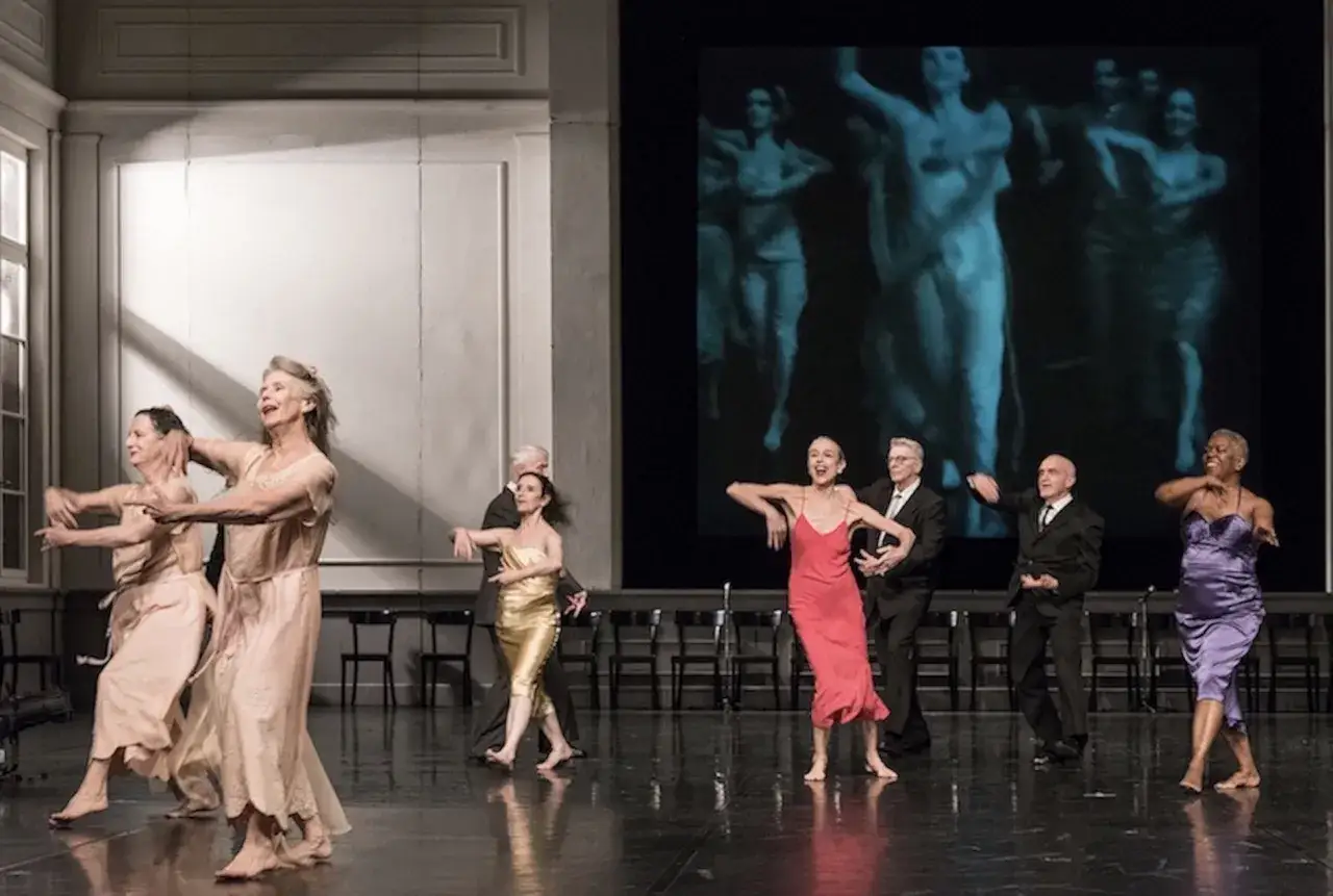 Ballerini in abiti leggeri e colorati danzano su un palco, con immagini di un film di Pina Bausch proiettate sullo sfondo.