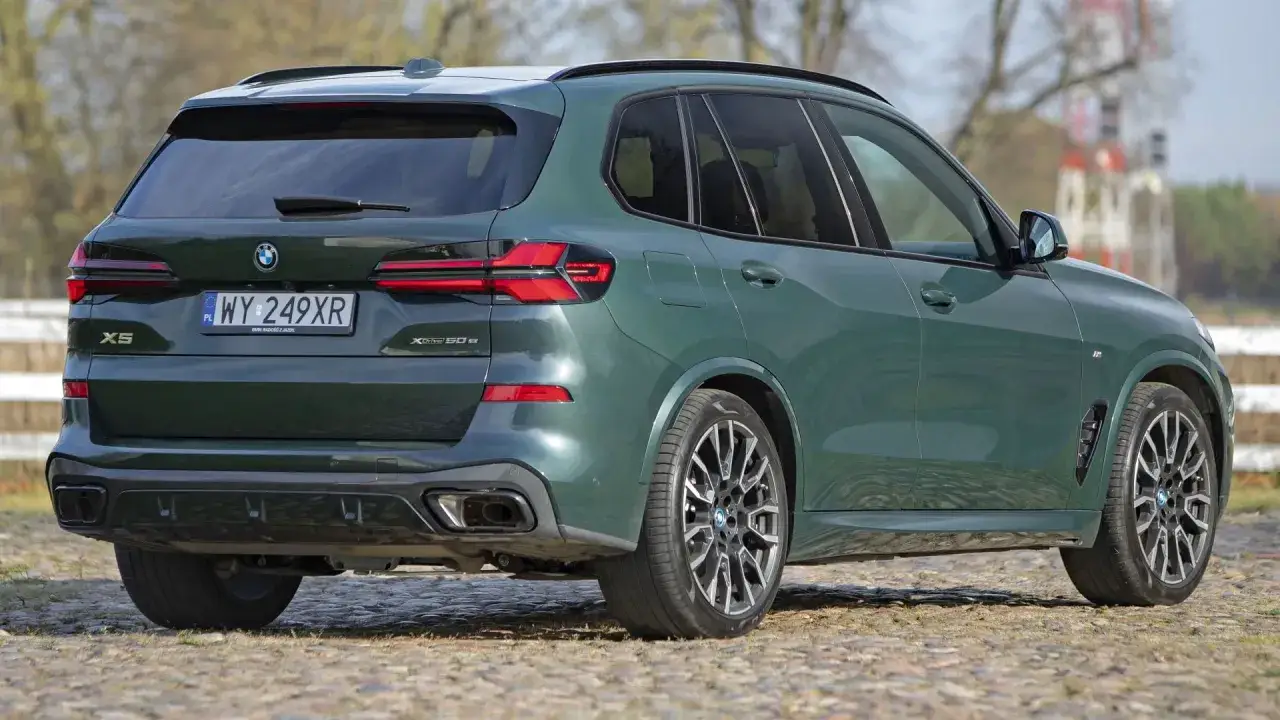 Nowe BMW X5 w kolorze ciemnej zieleni prezentuje się elegancko na tle natury. Widoczny tył pojazdu z charakterystycznymi światłami i felgami.