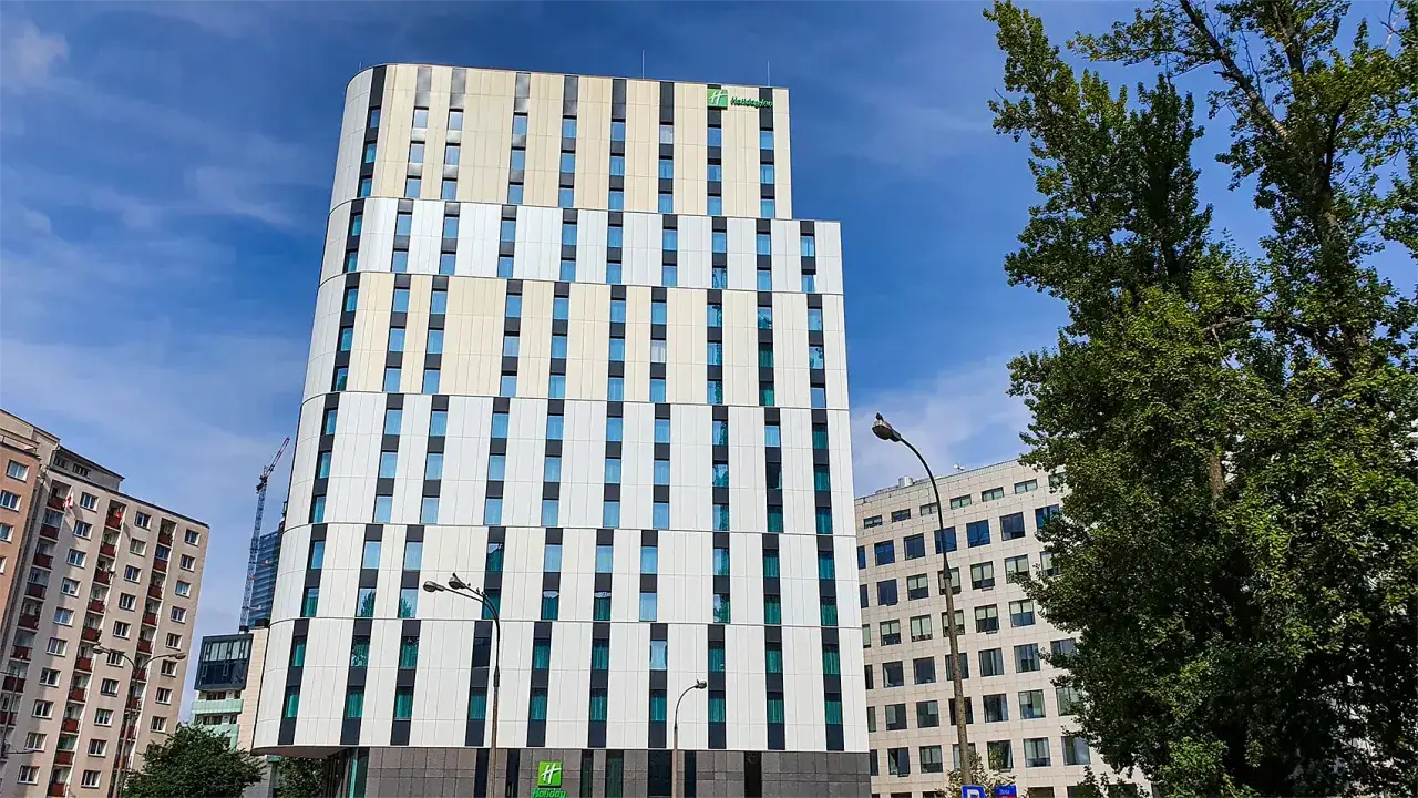 Nowoczesny budynek Holiday Inn Twarda w Warszawie, z charakterystyczną fasadą i niebieskimi oknami, na tle błękitnego nieba.