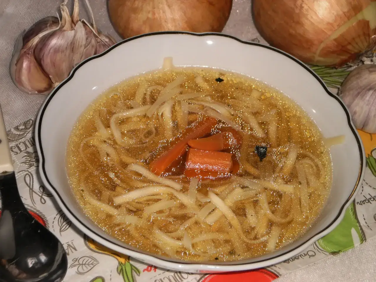 Aromaticzny rosół z indyka z makaronem i marchewką, podany w białej miseczce. Obok czosnek i cebula.