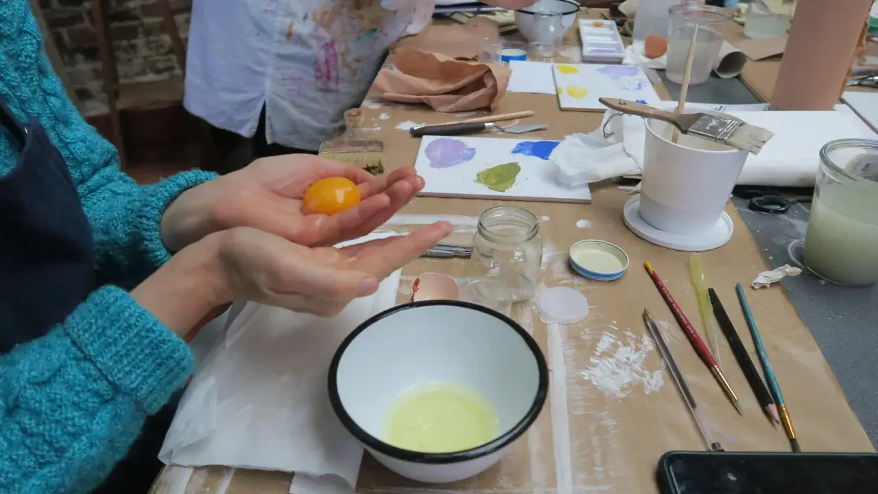 Pr&eacute;paration d'une peinture &agrave; l'&oelig;uf : une main tient un jaune d'&oelig;uf au-dessus d'un bol de blanc d'&oelig;uf, entour&eacute;e de mat&eacute;riel d'art.