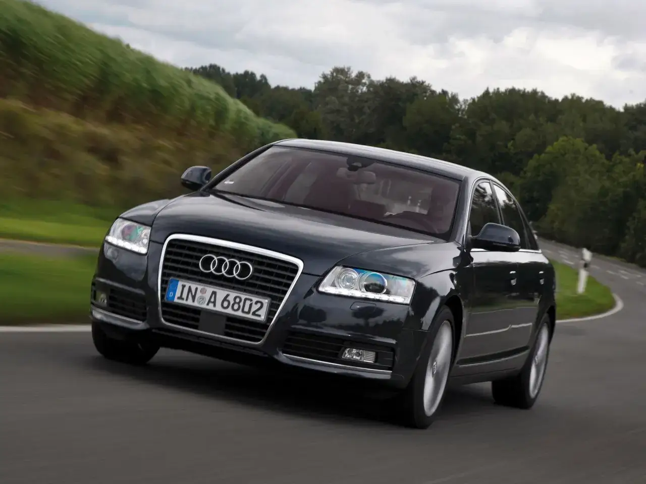 Ciemny Audi A6 C6 2.7 TDI zgrabnie pokonuje zakręt na malowniczej drodze.