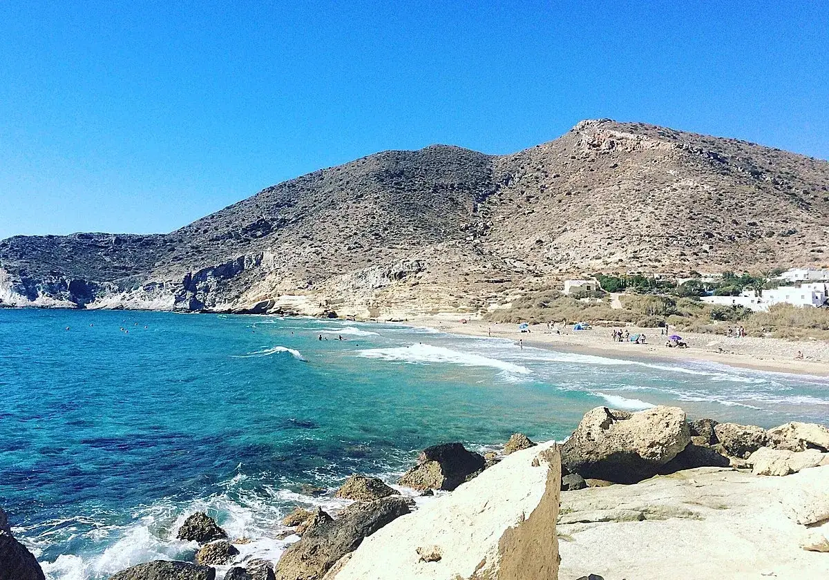 Aguas turquesas bañan la cala blanca de Almería, con montañas áridas de fondo y gente disfrutando del sol en la arena.