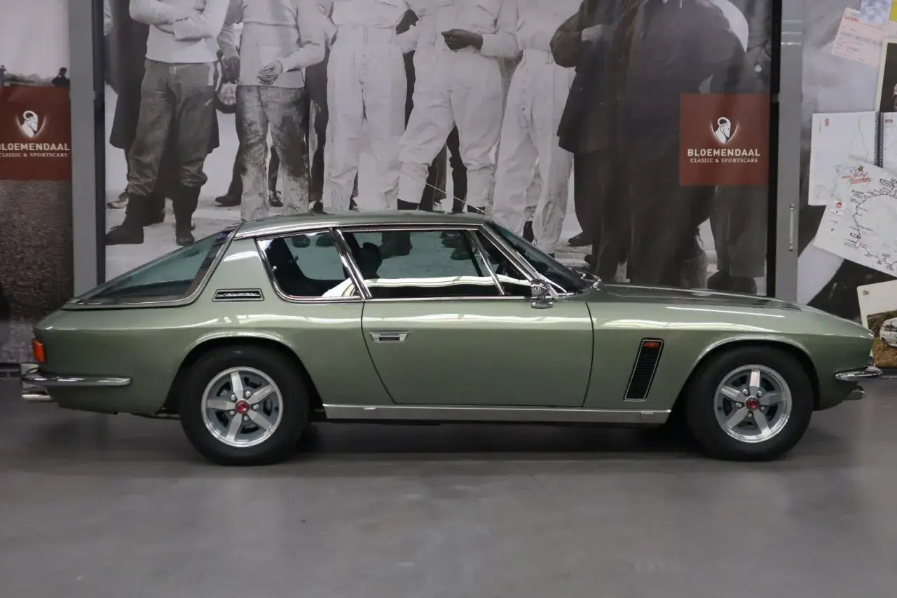 En gr&ouml;n Jensen Interceptor st&aring;r framf&ouml;r en v&auml;gg med svartvita bilder av m&auml;nniskor.