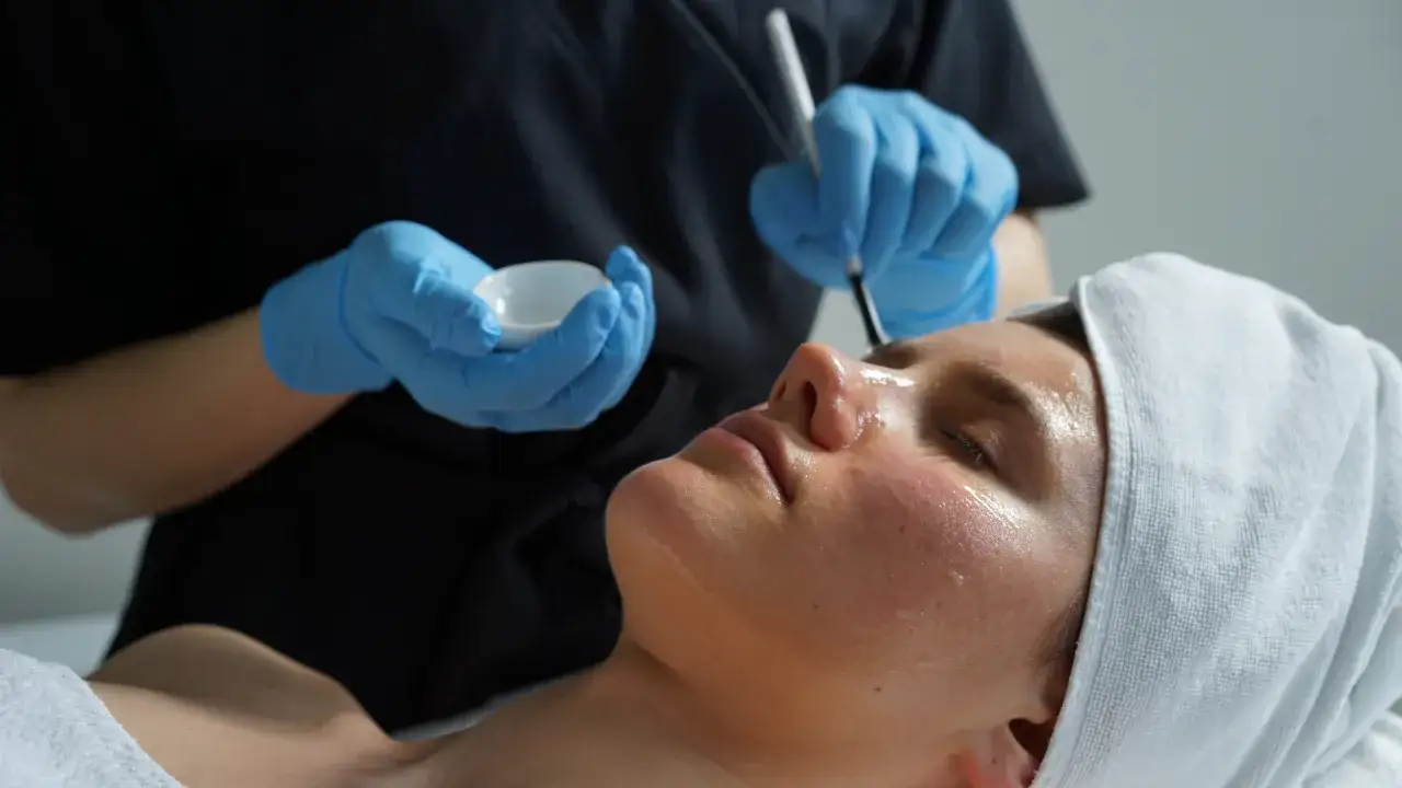 Kobieta z ręcznikiem na głowie przechodzi zabieg twarzy po peelingu chemicznym. Kosmetolog w rękawiczkach nakłada preparat.