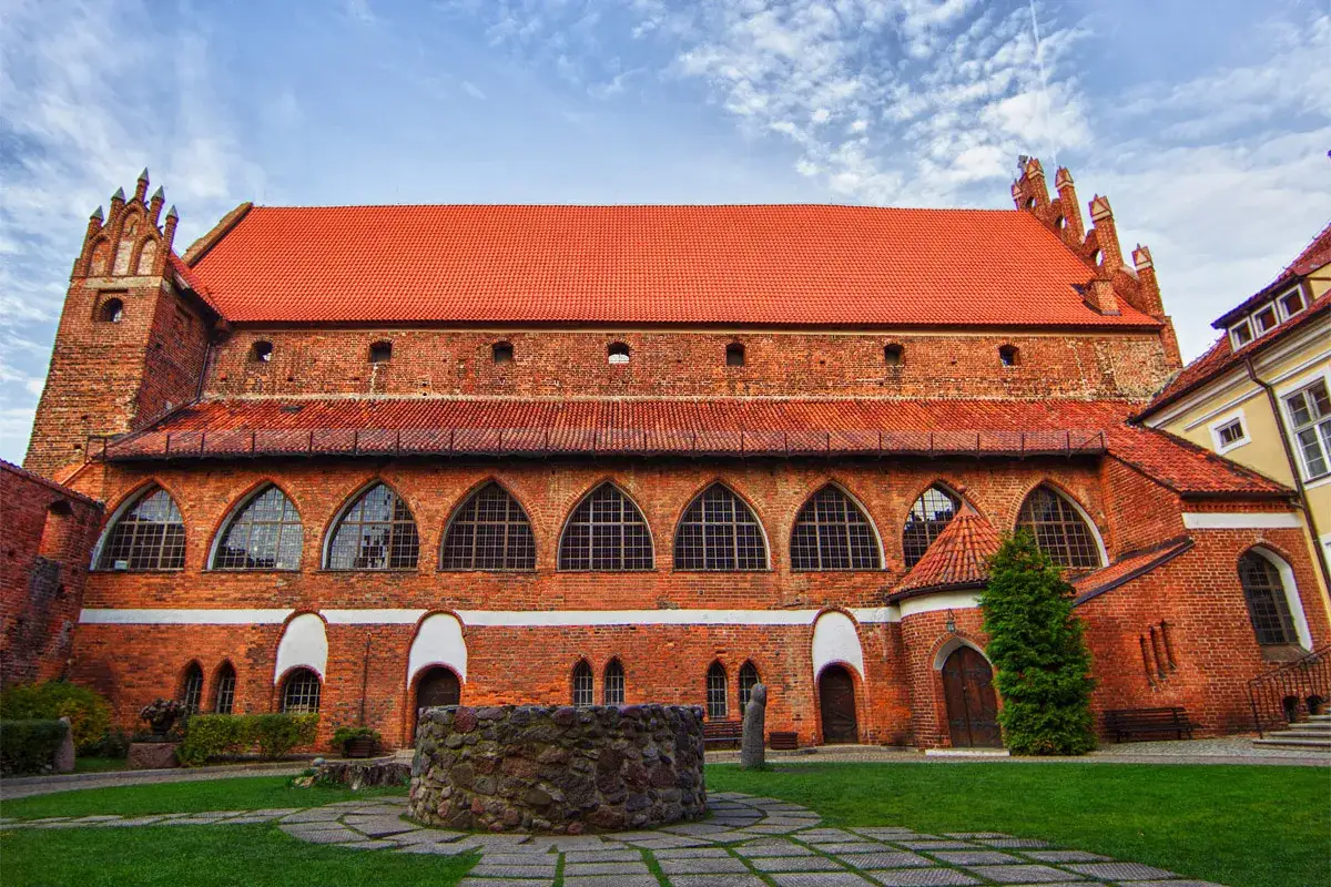Zamek kapituły warmińskiej z cegły, z charakterystycznym czerwonym dachem i łukowymi oknami, otoczony zielonym dziedzińcem z kamienną studnią.