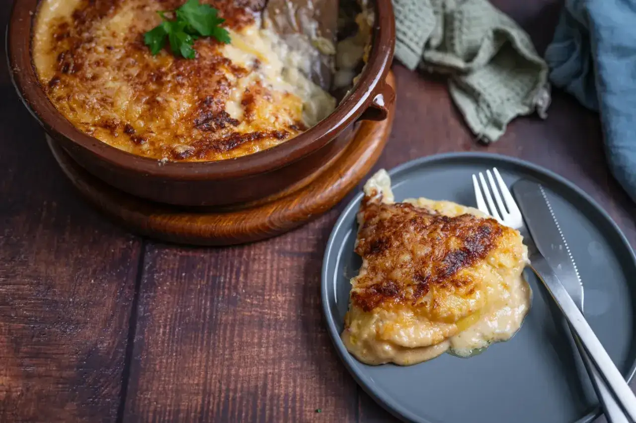 Ein goldbraun &uuml;berbackenes Kartoffelgratin-Rezept, serviert in einer Terrakotta-Sch&uuml;ssel und auf einem Teller mit Messer und Gabel.