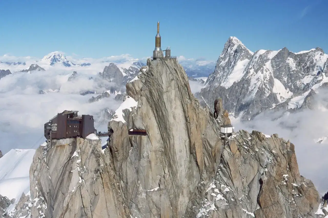 Widok na szczyt Aiguille du Midi, gdzie znajduje się kolejka na Mont Blanc. Budynki i wieża na skalistym wierzchołku, otoczone ośnieżonymi górami i chmurami.
