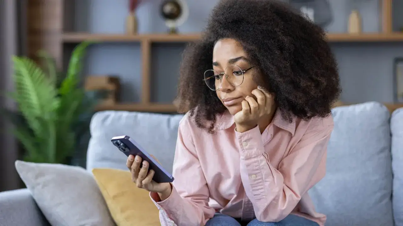 Ragazza con occhiali guarda il suo telefono, forse annoiata o pensierosa, mentre &egrave; seduta sul divano. La **second screen** cattura la sua attenzione.