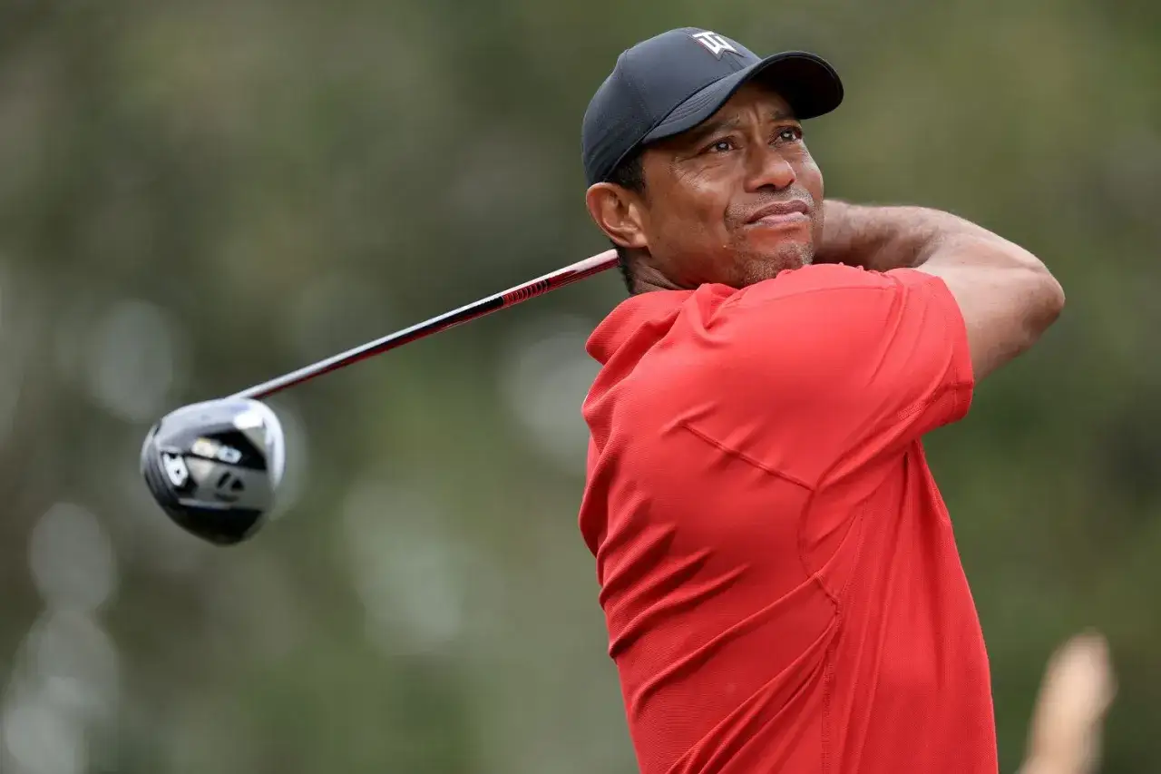 Tiger Woods w czerwonej koszulce i czapce z daszkiem wykonuje zamach kijem golfowym. Jego majątek to wynik lat sukces&oacute;w na polu golfowym.
