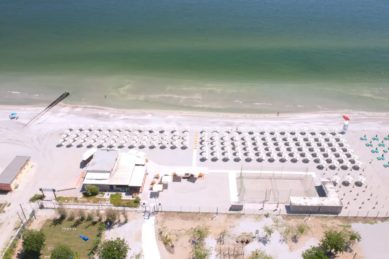 Lido di Savio, Milano Marittima: spiaggia ordinata con file di ombrelloni bianchi, mare calmo e una persona che cammina sull'acqua.