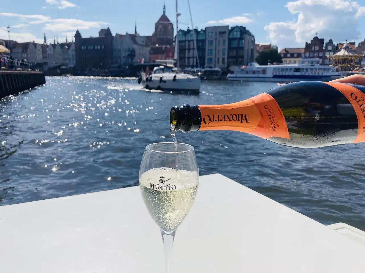 Prosecco Mionetto nalewane do kieliszka na tle wodnych widoków i zabytkowych budynków.