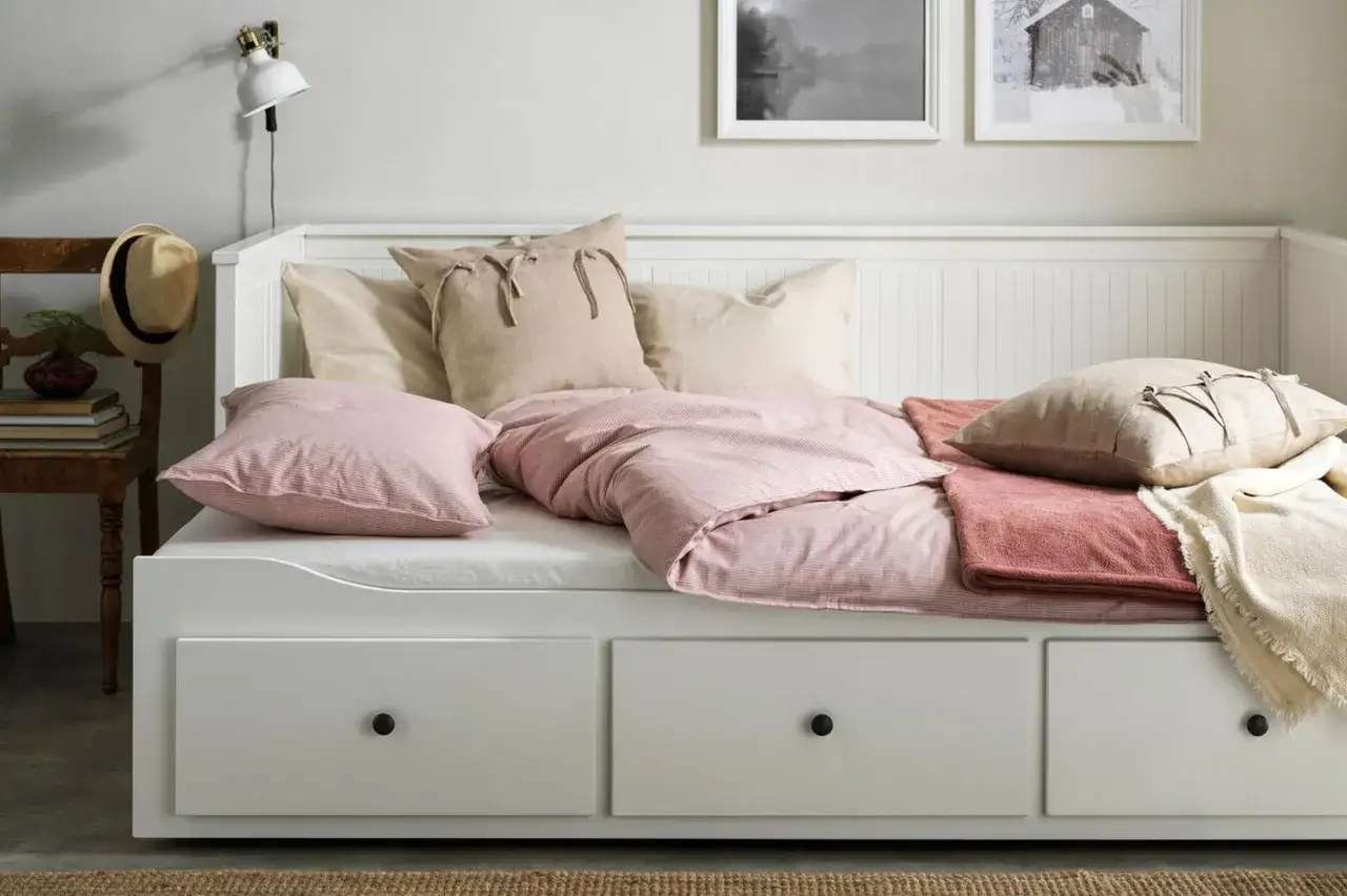Ein weißes Tagesbett mit Schubladen, das als Sofa gestaltet ist. Rosa und beige Kissen sowie Decken laden zum Entspannen ein.