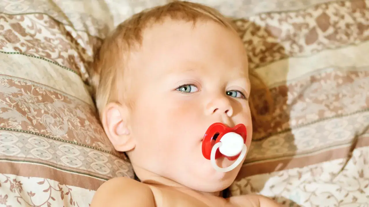 A baby with blue eyes and blonde hair lies on a patterned blanket, a red pacifier in their mouth. The pacifier has a slight pacifier bite mark.