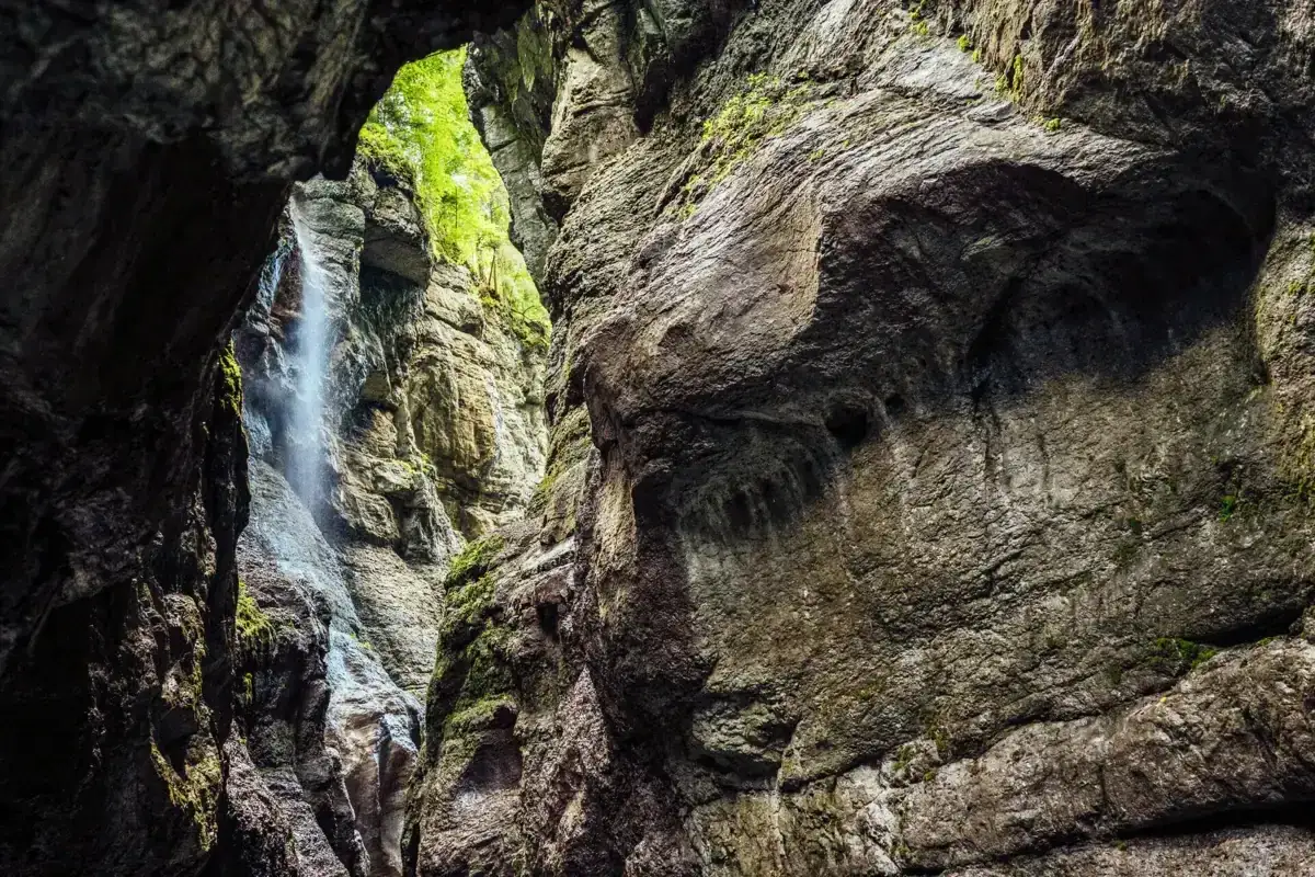 Tiefe Schlucht mit Wasserfall w&auml;hrend einer Partnachklamm Wanderung. Moosbewachsene Felsw&auml;nde ragen empor, dar&uuml;ber gr&uuml;nes Laub.