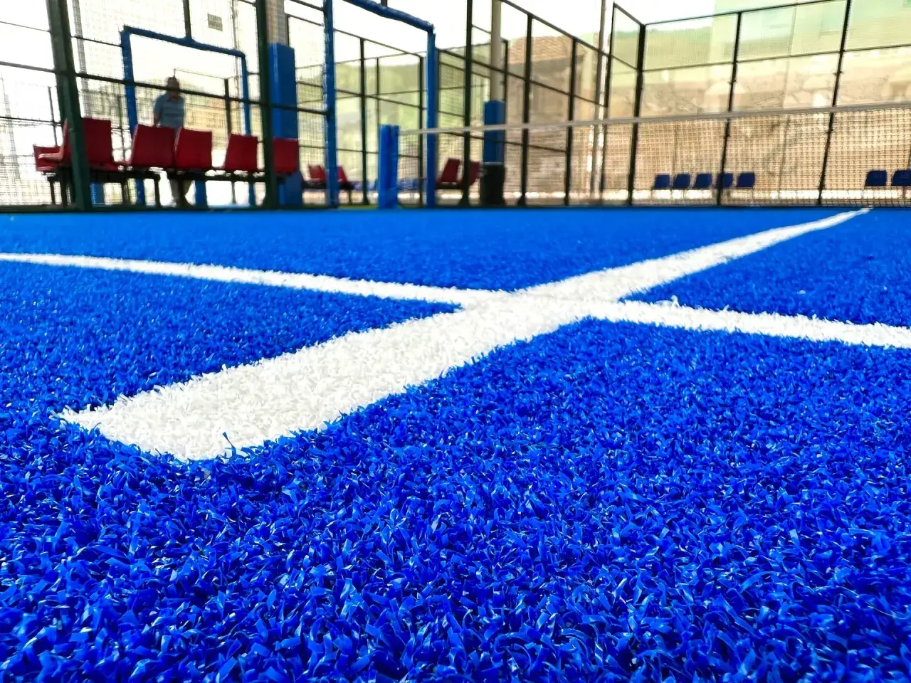 Cancha de p&aacute;del con c&eacute;sped artificial azul vibrante y l&iacute;neas blancas. Bancos rojos y una persona en el fondo.