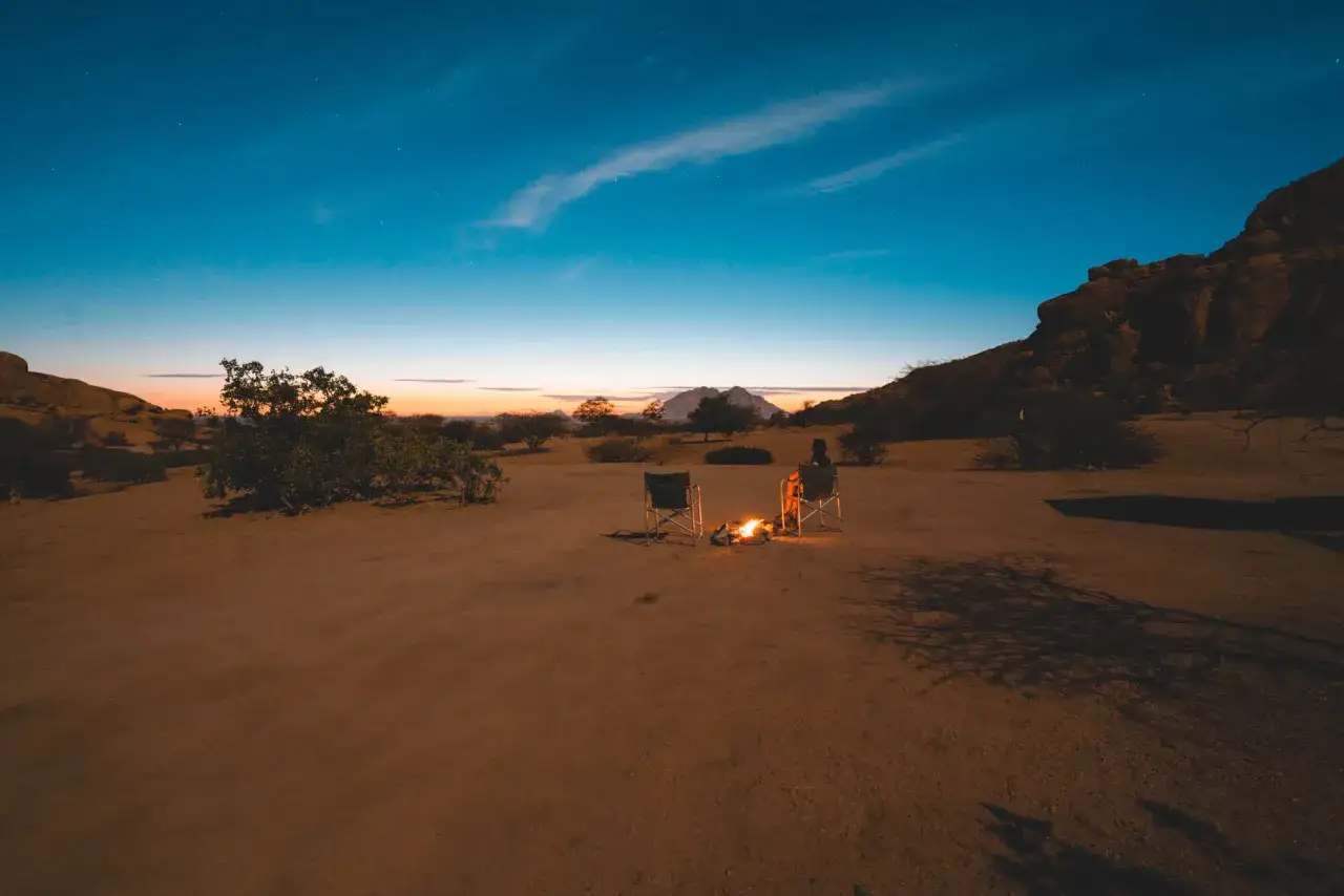 Abendstimmung bei den Spitzkoppe in Namibia. Ein Lagerfeuer wärmt zwei Stühle in der weiten Wüstenlandschaft unter dem Sternenhimmel.