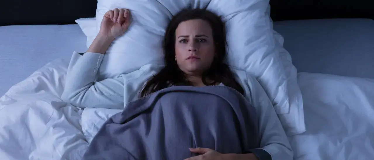 Frau liegt wach im Bett, Augen weit aufgerissen, die Decke fest umklammert. Ein schrecklicher Albtraum hält sie gefangen.