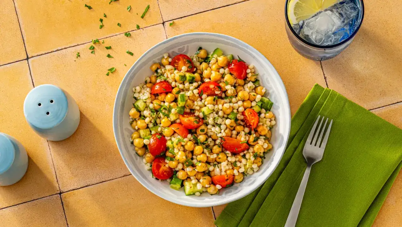 Frischer Couscous-Salat mit Kichererbsen, Tomaten und Gurken, garniert mit Petersilie. Daneben ein Glas Wasser mit Zitrone und Eis.