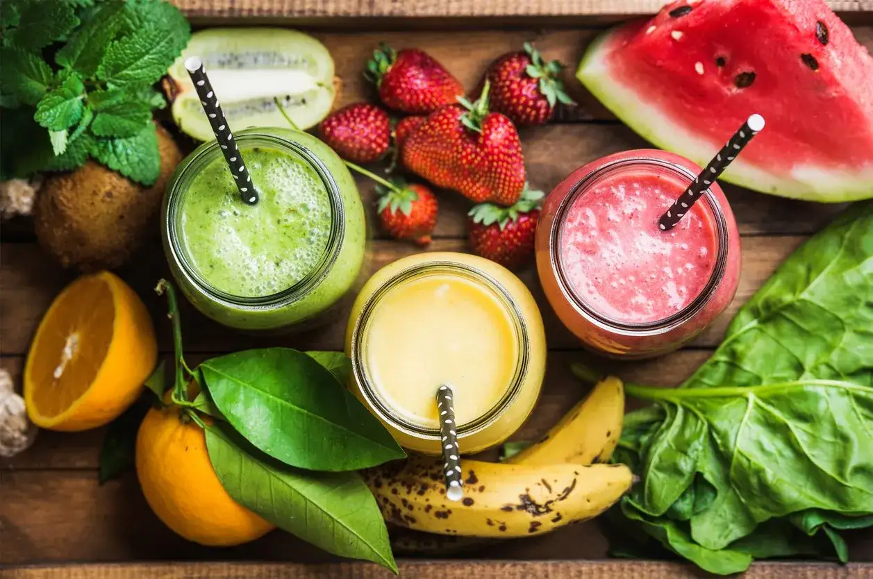 Kolorowe smoothie: zielone, żółte i różowe. Obok składniki: truskawki, kiwi, pomarańcze, banany, arbuz i szpinak. Idealne na lato, jak zrobić smoothie.