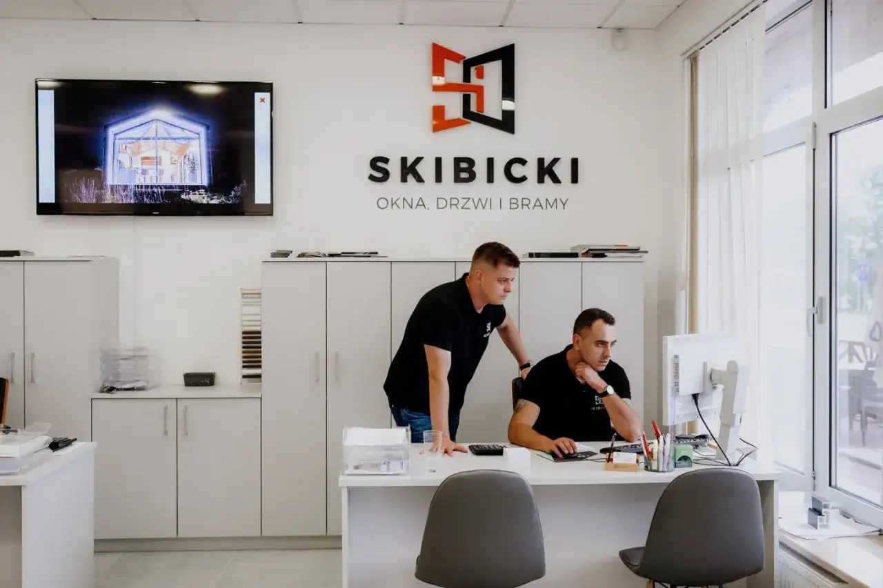 Dwóch mężczyzn w biurze firmy okiennej, jeden pracuje przy komputerze, drugi stoi obok.