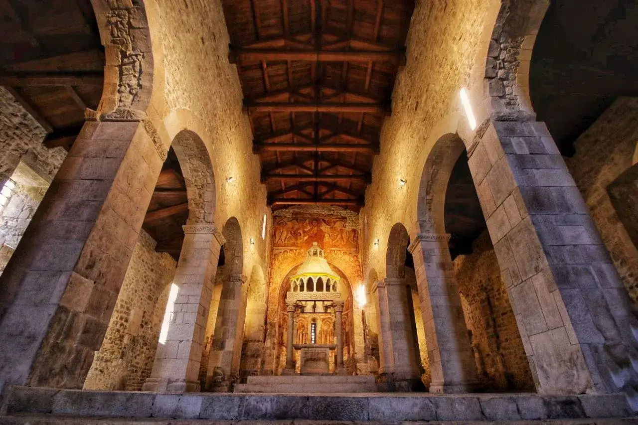 Interno di una chiesa romanica a Capestrano, cosa vedere: colonne massicce, archi in pietra, soffitto in legno e affreschi antichi sull'altare.