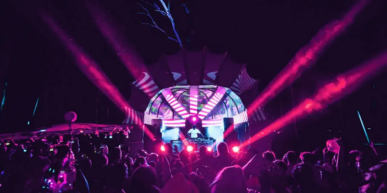 Un DJ mixa musica techno sotto un palco illuminato da luci rosa e viola, mentre una folla festeggia.
