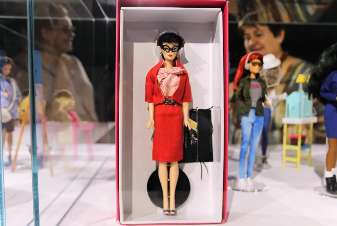 Lalka Barbie w czerwonym kostiumie, symbol emancypacji kobiet w lalce, prezentowana w pudełku. W tle inne lalki i wystawa.
