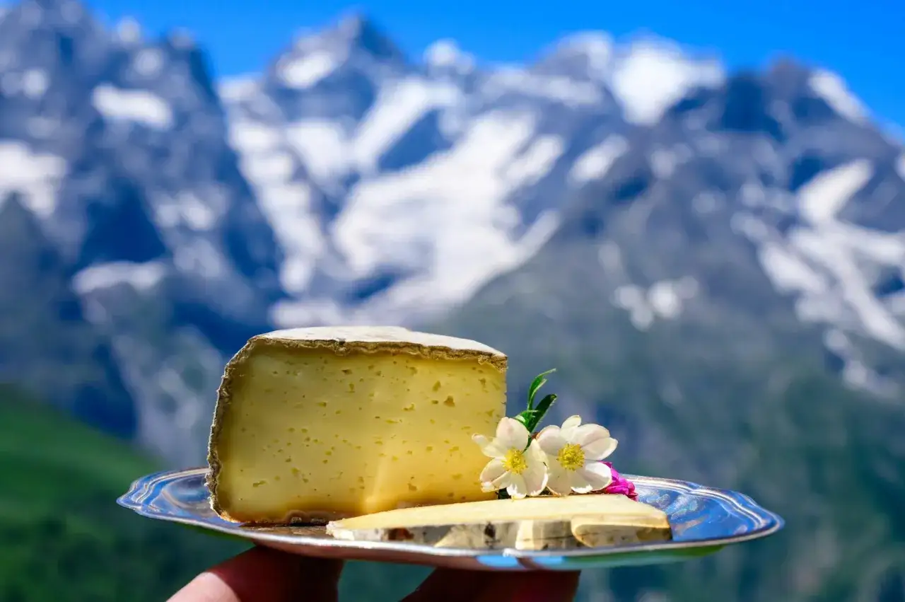 Un plateau de fromage savoyard, avec un gros morceau de fromage et une fine tranche, décoré de fleurs, sur fond de montagnes enneigées.