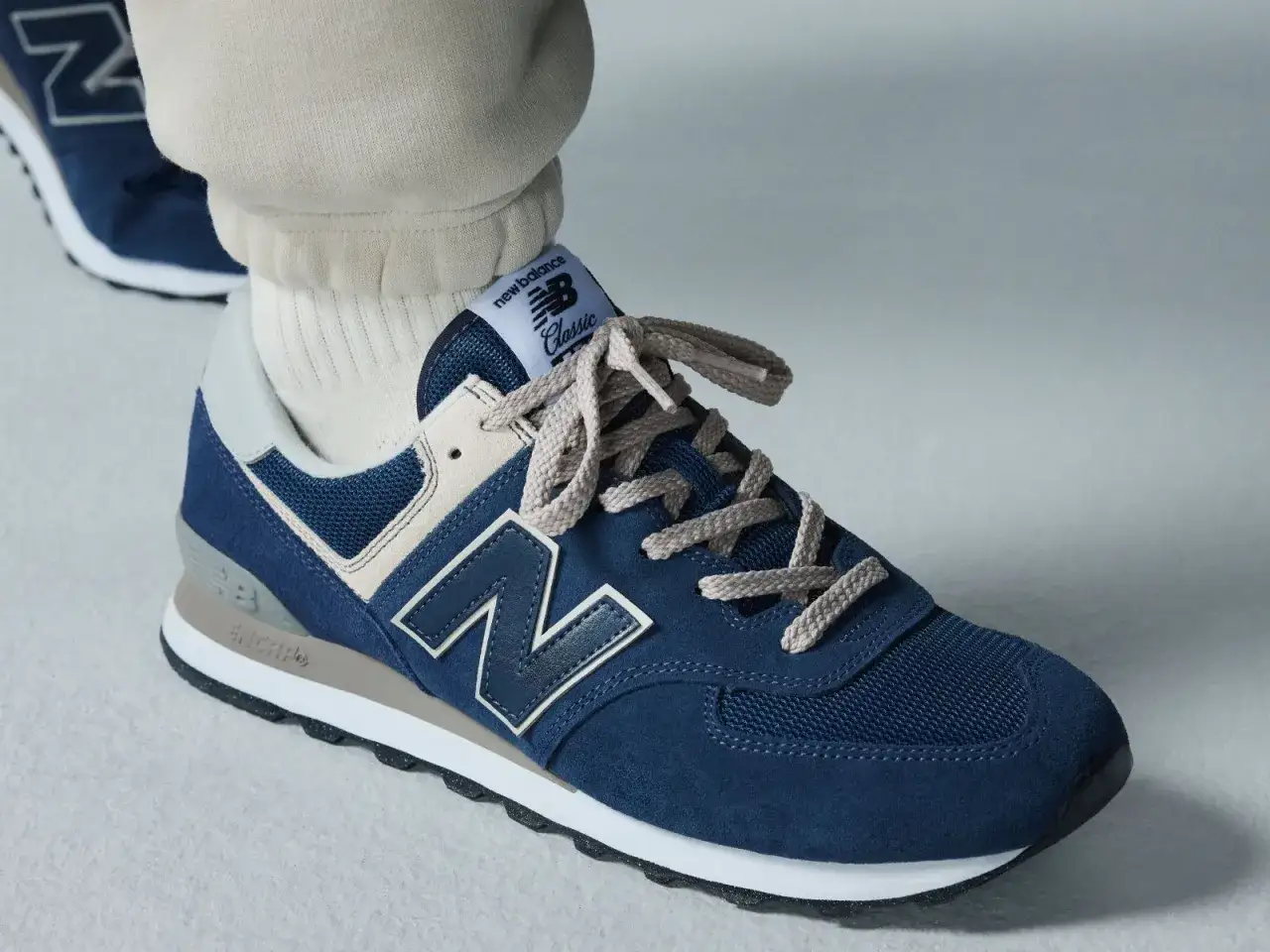Granatowe buty sportowe new balance wl 574 z beżowymi i szarymi akcentami, na stopie w beżowych spodniach dresowych i białych skarpetkach.