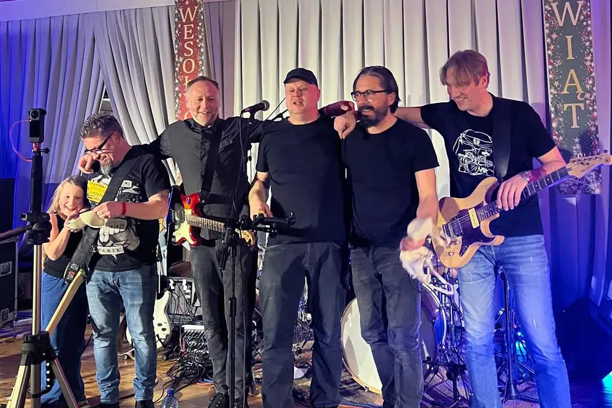 Zespół muzyczny na scenie, grający koncert. W tle wiszą dekoracje z napisem 