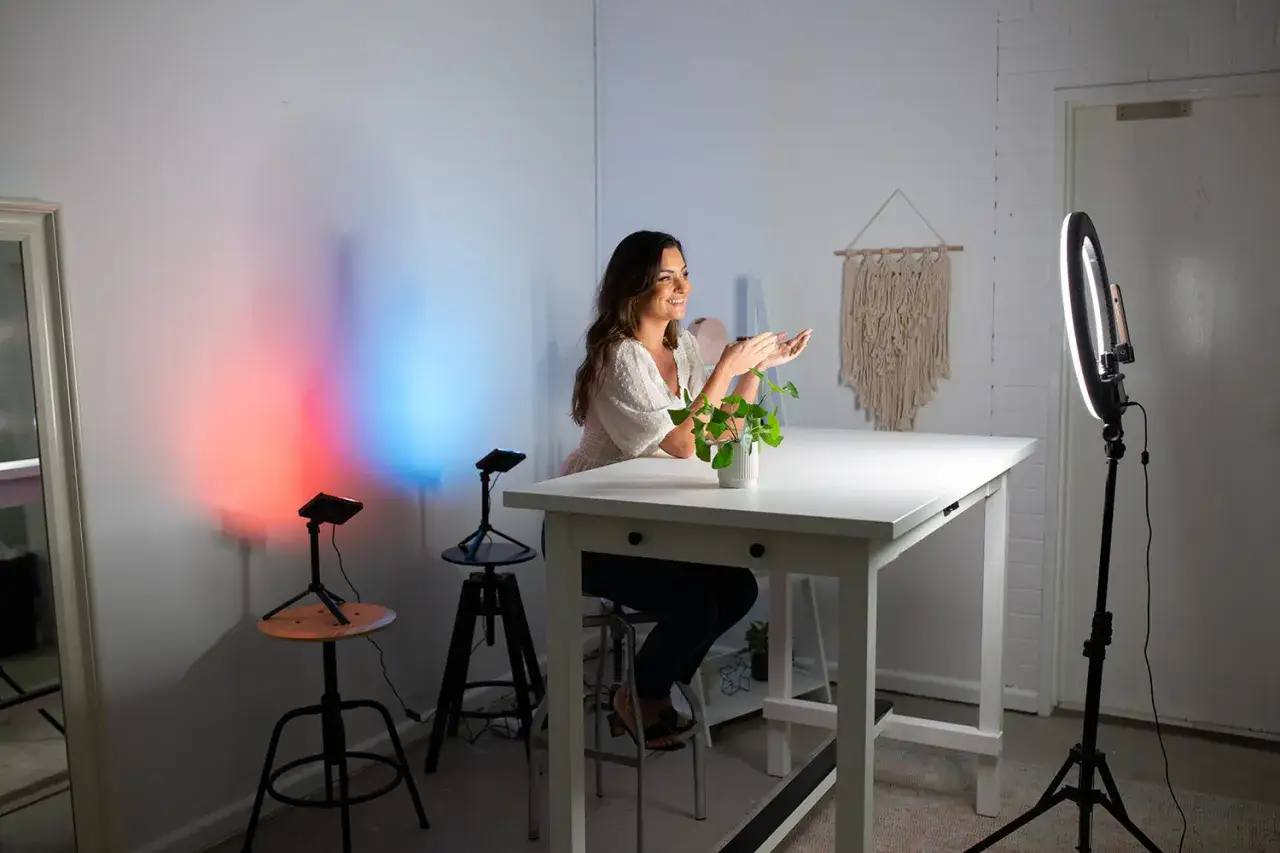 Uśmiechnięta kobieta przy stole, gotowa do nagrywania na YT. W tle kolorowe światła i ring light.