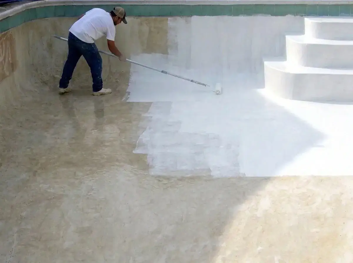 Hombre pintando una piscina de cemento con rodillo. La mitad de la piscina est&aacute; pintada de blanco, la otra mitad a&uacute;n est&aacute; en proceso.