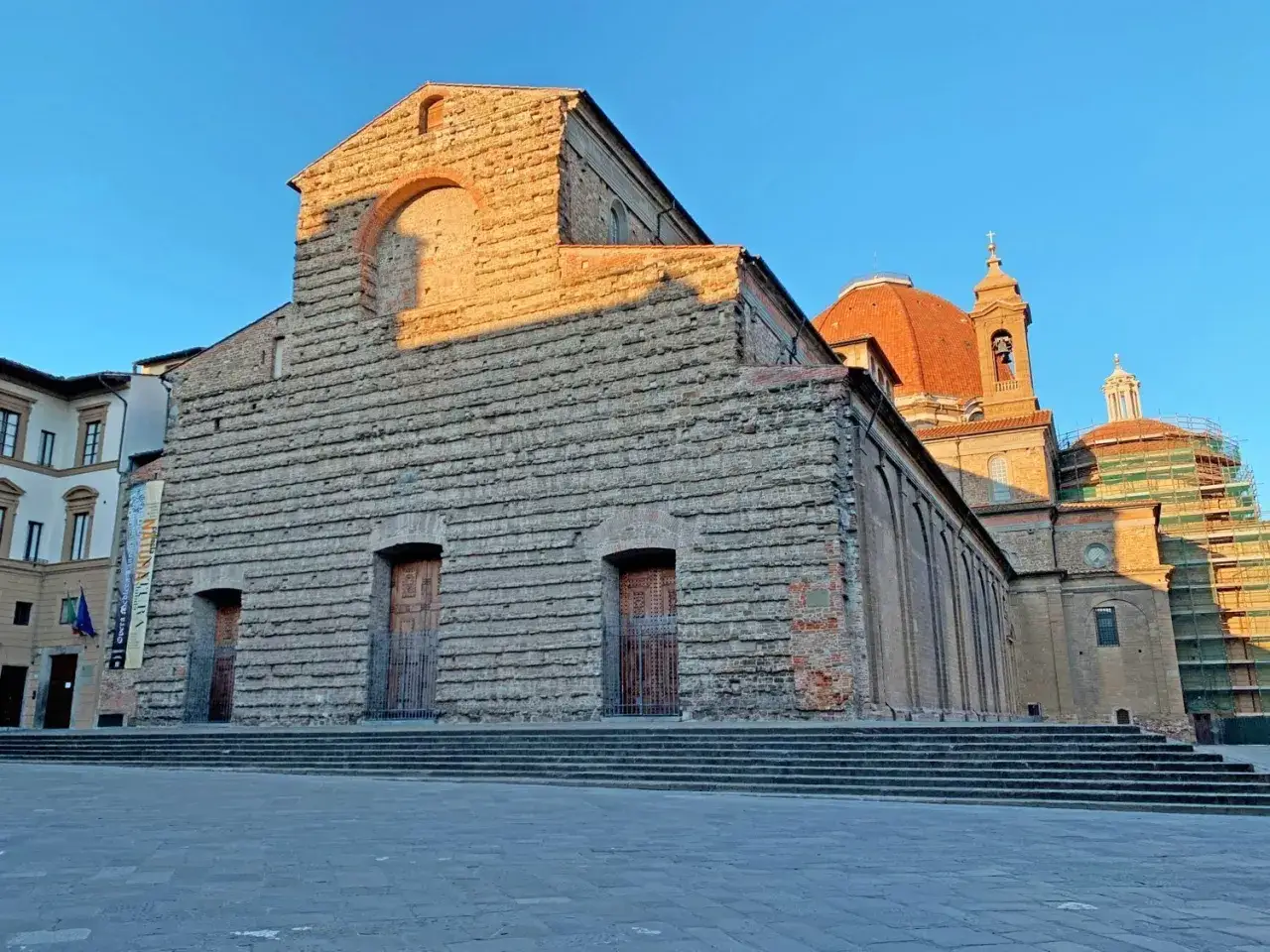 La facciata in pietra della basilica San Lorenzo, con le sue porte in legno e le gradinate, si staglia contro il cielo azzurro.